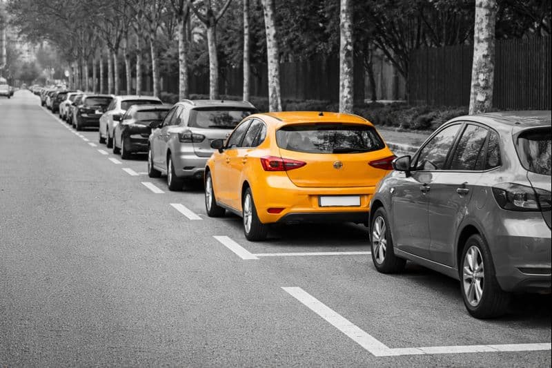 Fotografía urbana donde se ve un coche naranja estacionado correctamente entre otros vehículos en una fila de aparcamiento, destacando visualmente la importancia de controlar el espacio y las referencias al aparcar en bateria