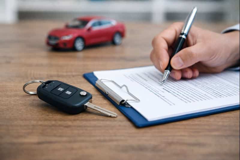 Imagen que representa un contrato de compraventa de coche aePlusTest, con una mano firmando un documento sobre una mesa, las llaves del vehículo en primer plano y un coche rojo de juguete al fondo, simbolizando el acuerdo legal entre comprador y vendedor