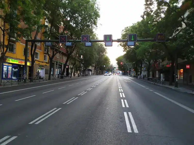 Avenida urbana amplia con señalización luminosa superior que regula un carril reversible mediante flechas verdes y aspas rojas sobre varios carriles